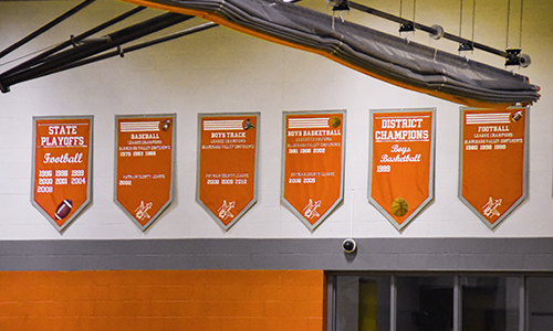 Six orange championship banners with white text hanging in a gymnasium.